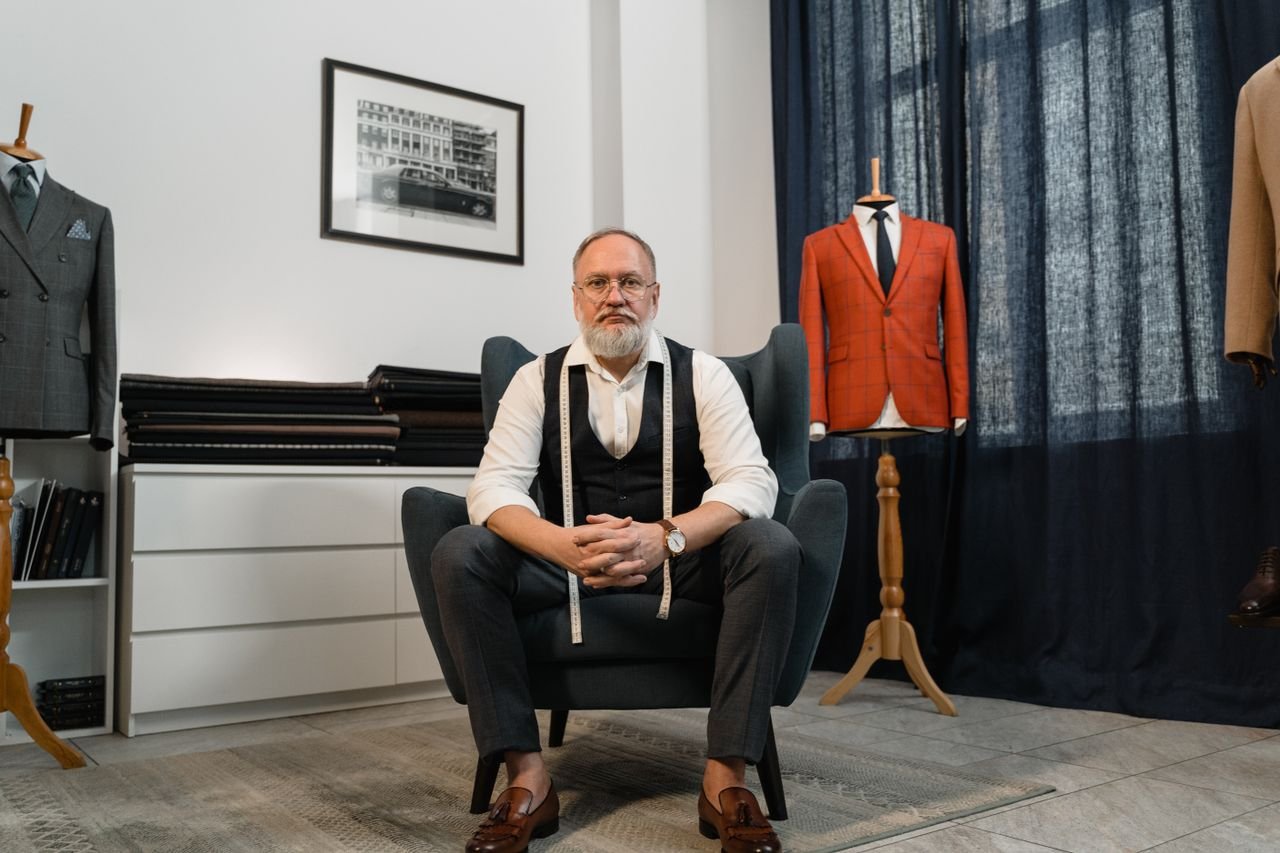 Elderly tailor sitting in an elegant shop with suits and mannequins.
