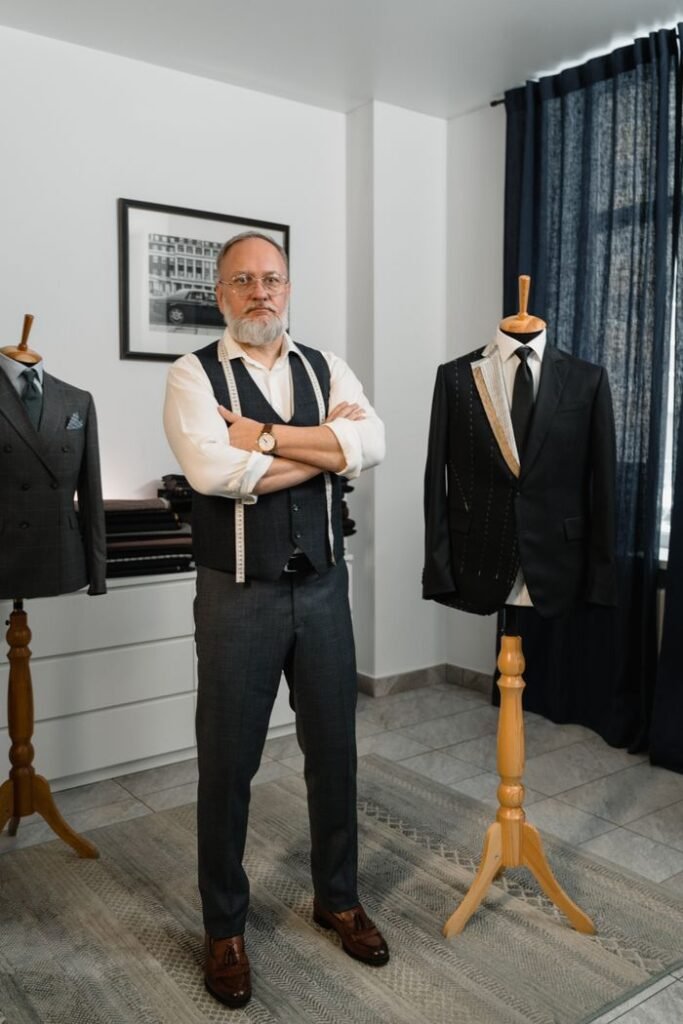 Tailor standing confidently in his studio with bespoke suits on mannequins.